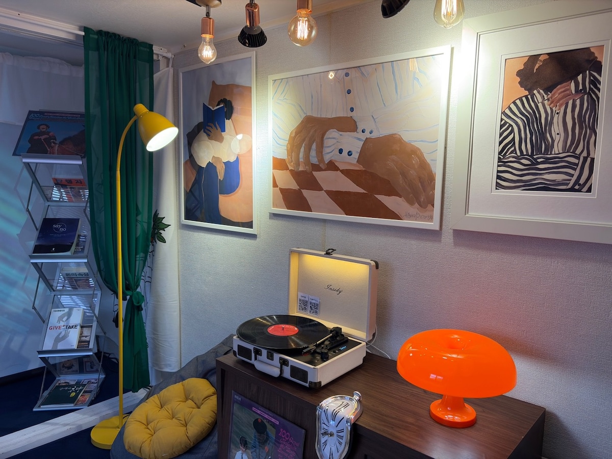 A cozy corner of the living area features a vintage record player atop a wooden table, surrounded by vibrant wall art. A warm light fixture hangs above, and a distinctive orange lamp adds a pop of color. A comfortable yellow pouf provides additional seating.