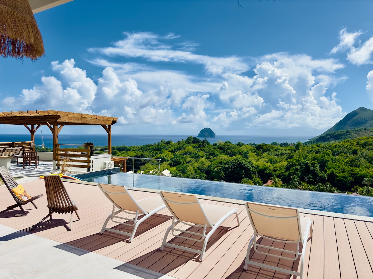 An infinity pool overlooks lush greenery and stunning ocean views, including the iconic Rocher du Diamant. Lounge chairs are positioned around the wooden deck, inviting relaxation. A shaded pergola provides additional outdoor seating, set against a backdrop of blue skies and white clouds.