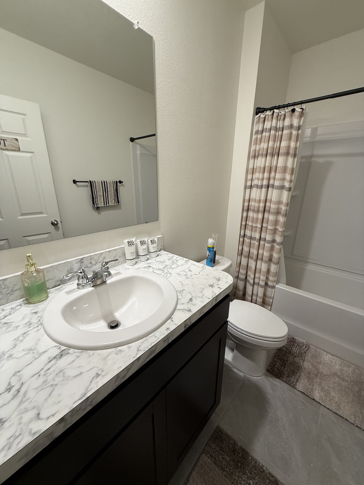 A clean bathroom features a marble countertop with a sink and mirror. Fresh towels are neatly arranged, and toiletries are provided. A shower curtain in a subtle pattern complements the space, while a toilet is located nearby, enhancing the simplicity and functionality of the layout.