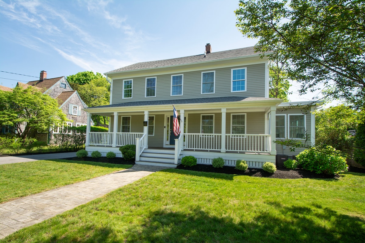 A two-story home is viewed from the front, featuring a wide porch with white railings. A flag hangs near the entrance. Well-maintained greenery surrounds the house, while a stone pathway leads to the front steps. The sky is clear, and natural light illuminates the exterior.