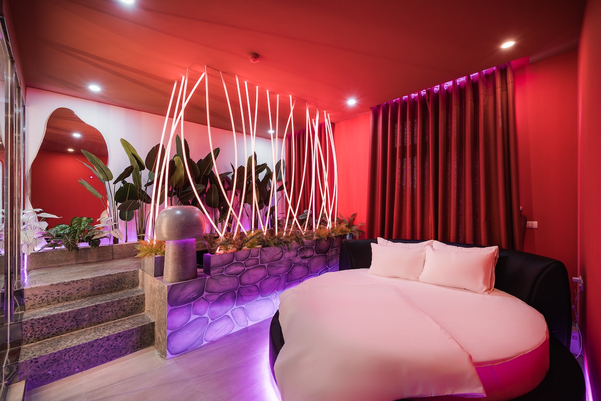 An inviting space features a round bed with soft pillows. Vibrant red walls are complemented by lush greenery and modern lighting, creating a unique atmosphere. Decorative plants are showcased, while soft ambient lights highlight the contours of the room, enhancing its contemporary design.