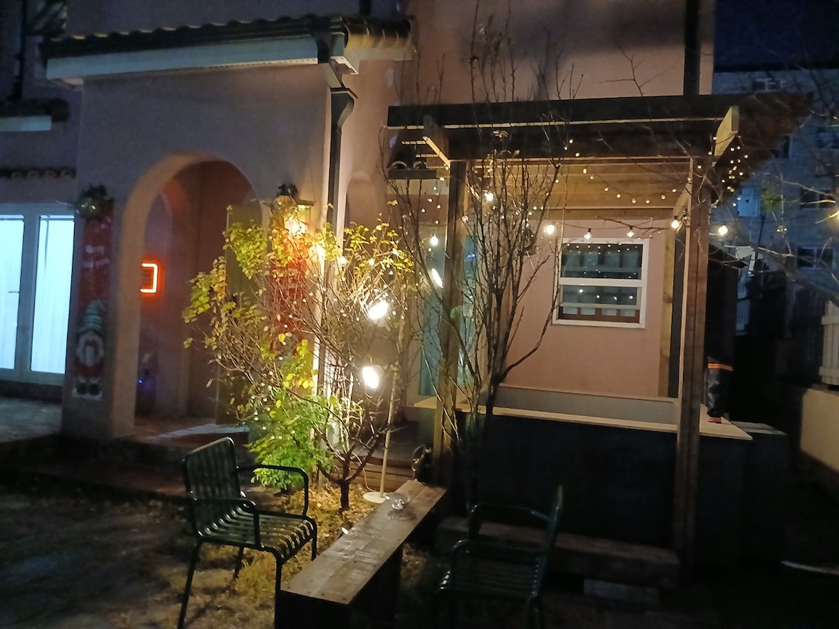 An outdoor seating area is depicted, featuring a wooden bench and two green chairs. String lights illuminate the scene while trees with bare branches provide a natural touch. The warm glow from a window hints at cozy interior spaces nearby.