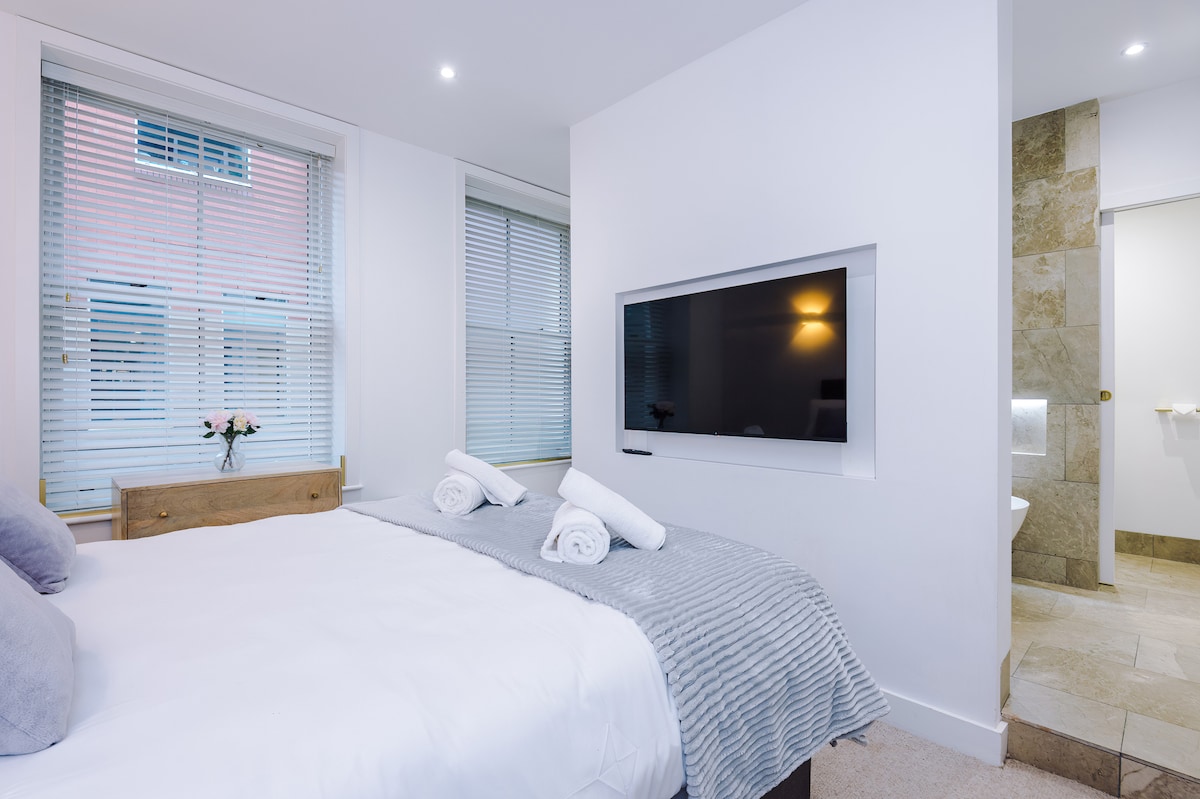 A master bedroom features a spacious bed adorned with soft linens, flanked by two rolled towels at the foot. A wall-mounted TV is set opposite the bed, with natural light filtering through the nearby window covered by blinds. A private en-suite bathroom is visible.