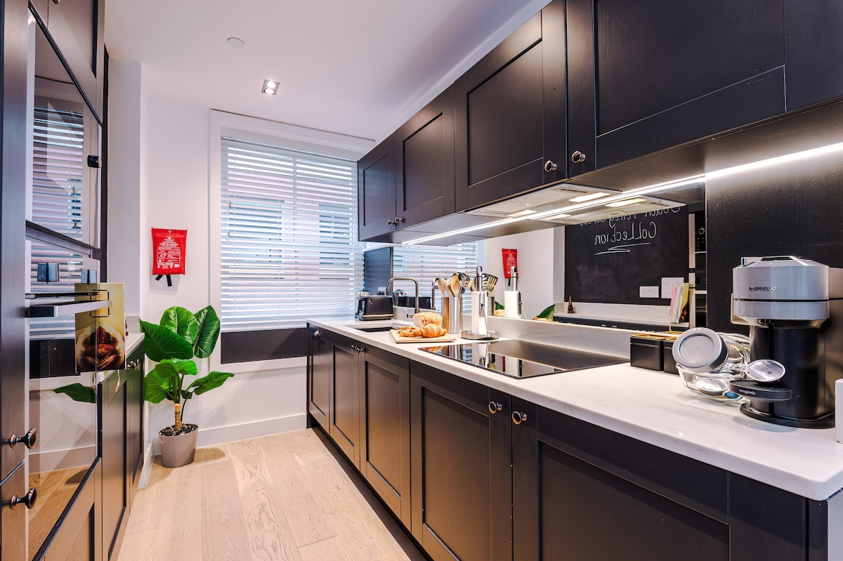 A modern kitchen area is presented with dark cabinetry and sleek countertops. Appliances including a coffee maker and an integrated stovetop are visible. Natural light enters through layered blinds, while a small potted plant adds a touch of greenery to the space.