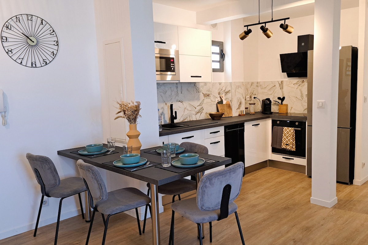 A modern open-plan kitchen and dining area is visible, featuring sleek black countertops and white cabinetry. A table set for four is adorned with blue plates and glassware. The space is complemented by stylish light fixtures and a wall clock, enhancing the overall ambiance.