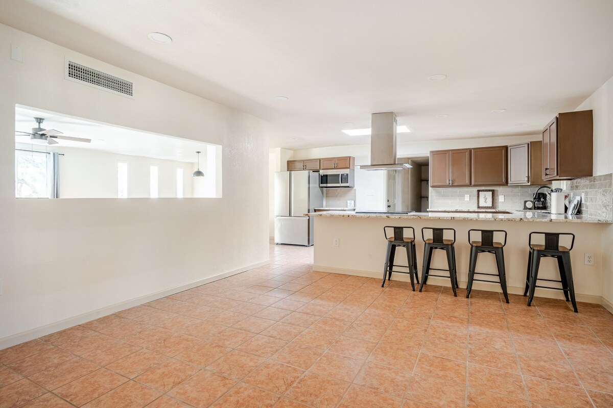 An open-concept kitchen and dining area features a large island with four black metal bar stools. The kitchen is equipped with stainless steel appliances and dark cabinetry, creating a modern look. Light beige walls and tiled flooring contribute to a bright and spacious atmosphere.