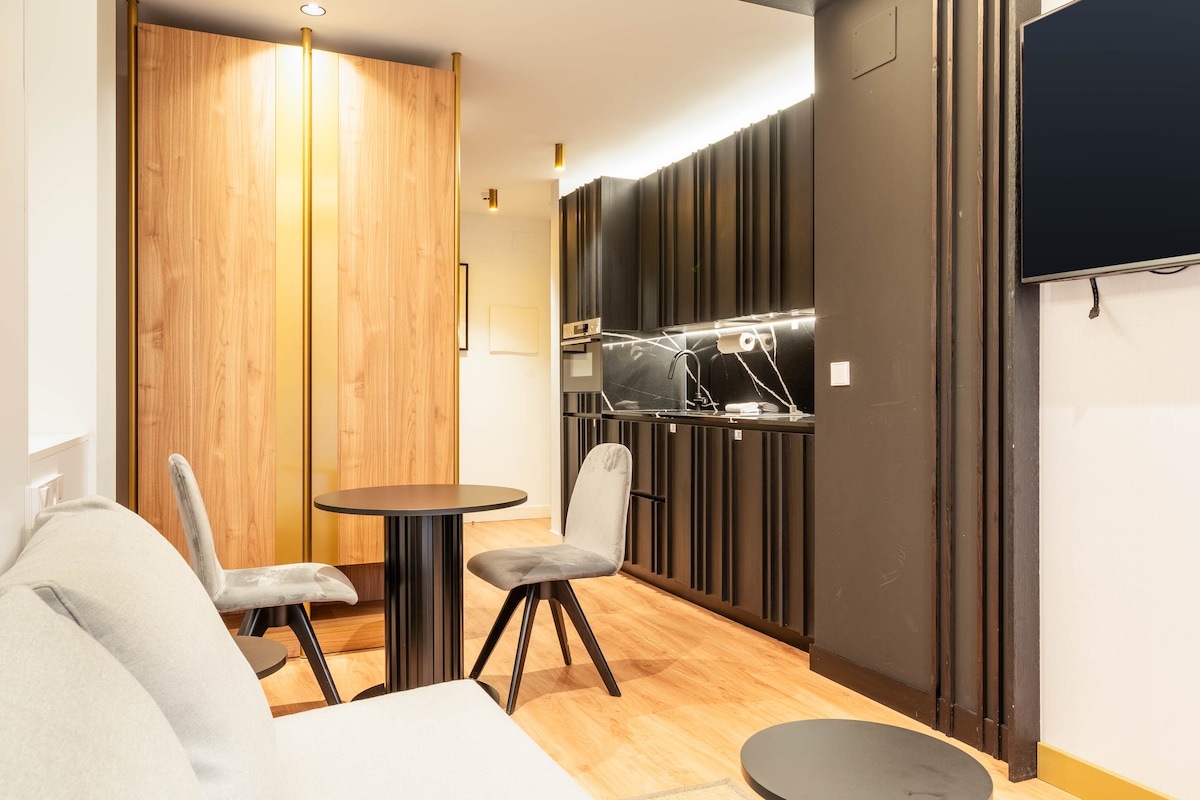 A modern living area showcases a stylish, neutral-colored sofa opposite a round dining table accompanied by two chairs. To the right, a sleek kitchen features dark cabinetry and well-integrated appliances, complemented by warm wood accents. A wall-mounted TV is visible for entertainment.