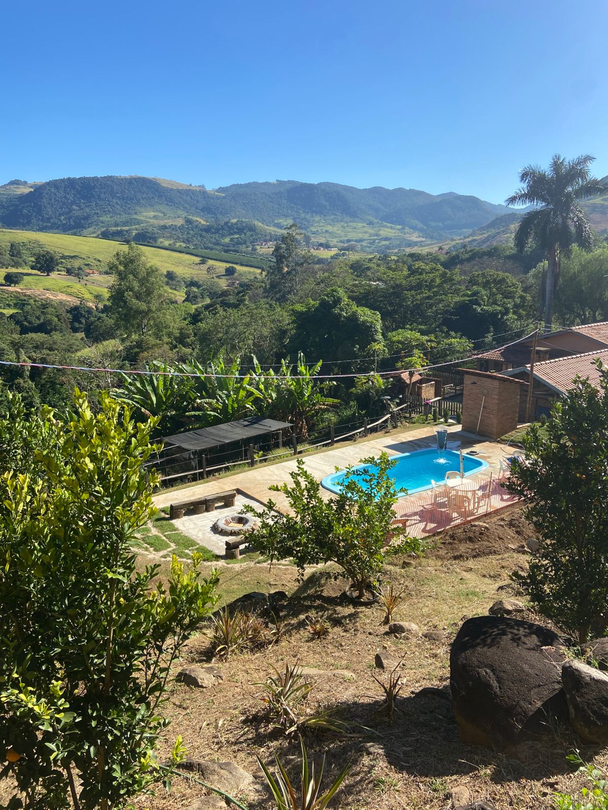 A scenic view of a pool area surrounded by lush greenery and rolling hills. The pool area features lounge chairs and a shared fire pit, while a rustic brick house is visible in the background, integrating harmoniously with the natural landscape.