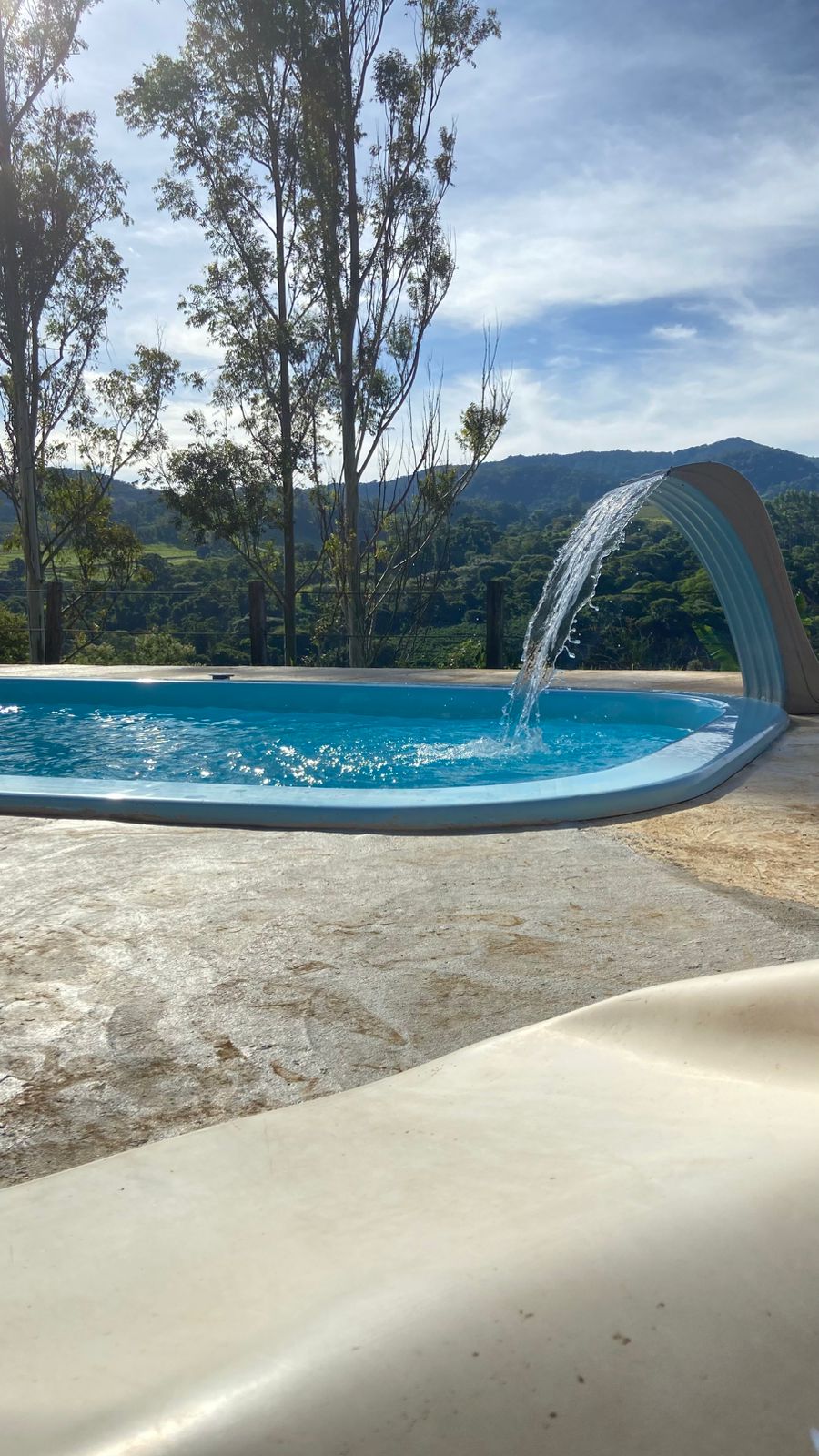The swimming pool features a gentle waterfall cascading into the clear blue water, surrounded by a spacious deck. Tall trees provide shade in the background, and the scenic hillside can be seen beyond the pool area, enhancing the tranquil environment.