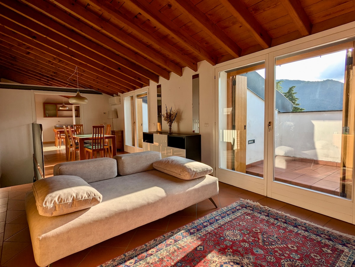 A spacious living area is highlighted by a large, light-colored sofa and a decorative rug. Expansive glass doors allow natural light to fill the space, leading to a terrace. The ceiling features exposed wooden beams, adding character to the inviting environment.