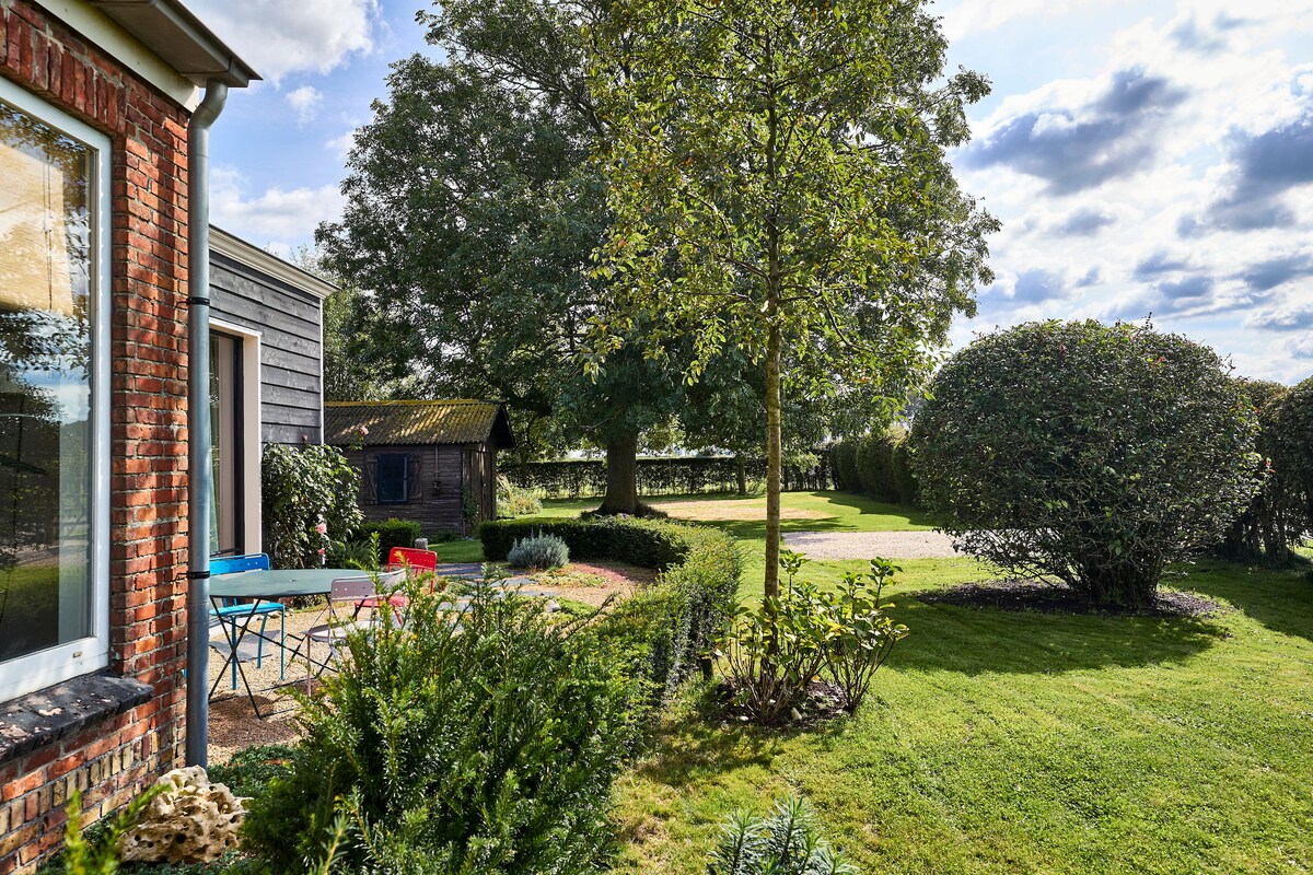 A spacious garden area is surrounded by neatly trimmed hedges and features a large tree providing shade. Colorful outdoor furniture is arranged on a gravel path, inviting relaxation. A wooden shed is visible in the background, nestled among vibrant greenery under a partly cloudy sky.