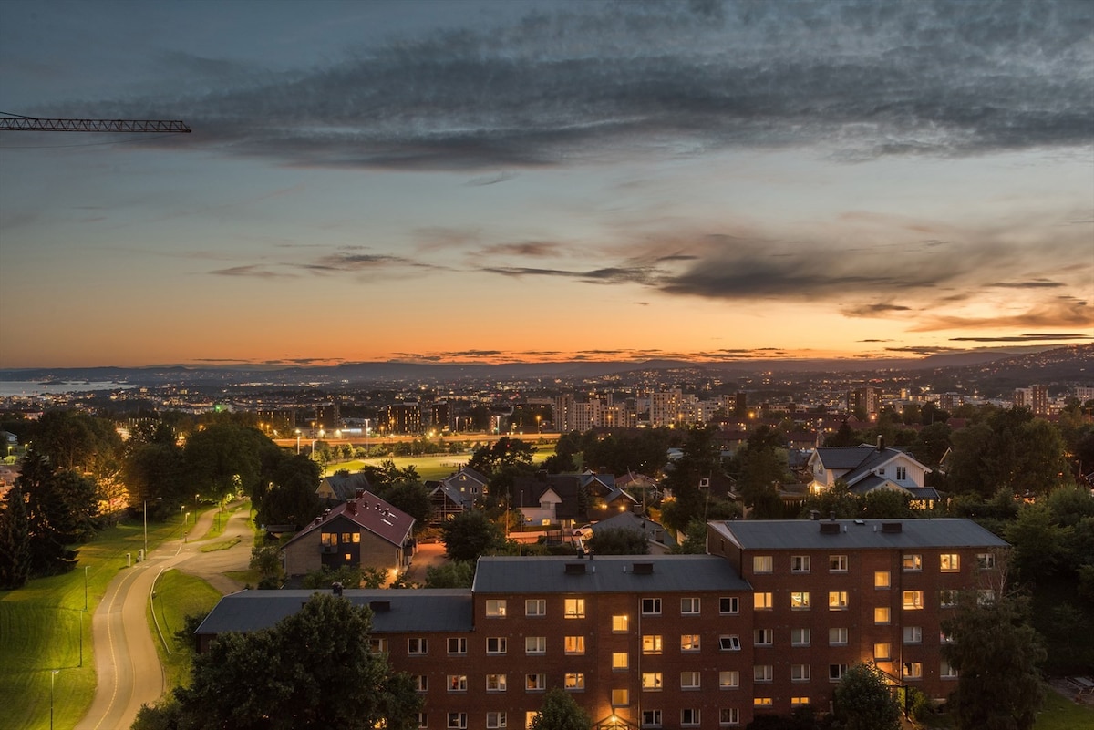 A panoramic view of Oslo during sunset is presented, showcasing the cityscape along with its illuminated buildings and streets. The horizon features a gradient of warm colors as the sun sets, with greenery and residential areas visible in the foreground.