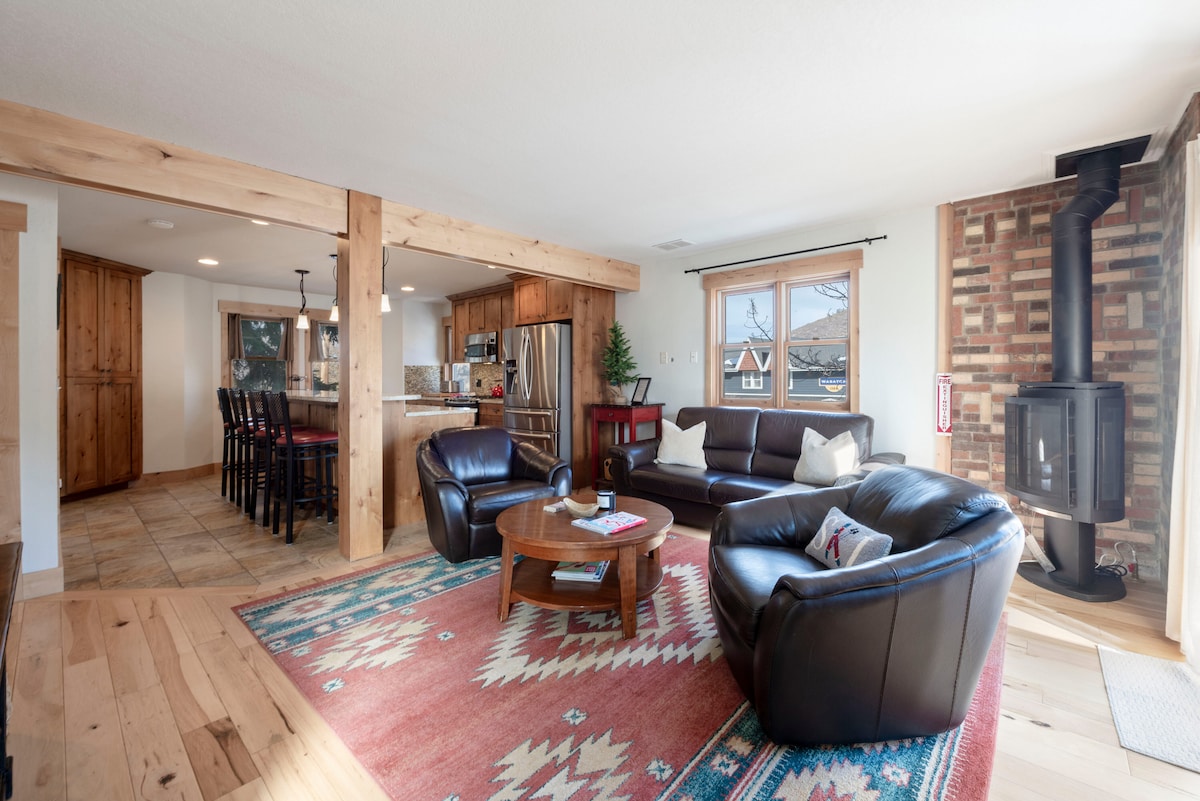 A welcoming living area features a comfortable seating arrangement, including a spacious dark leather sofa and two chairs. A wooden coffee table is centrally placed on a patterned rug. A modern stove is visible beside a stone wall, with light streaming in through large windows.