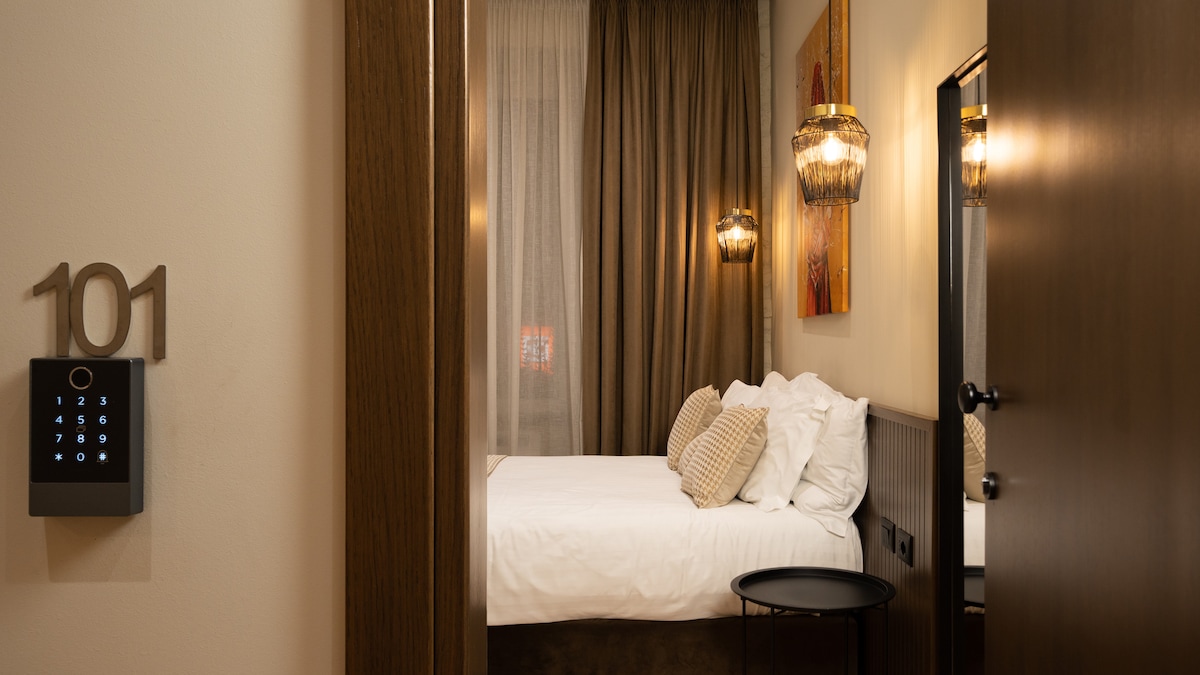 The entryway reveals a chic boutique room, featuring a spacious bed with decorative pillows. Soft curtains frame the window, and ambient lighting is provided by stylish fixtures. A small side table complements the modern decor accented by wall art, creating an inviting atmosphere.
