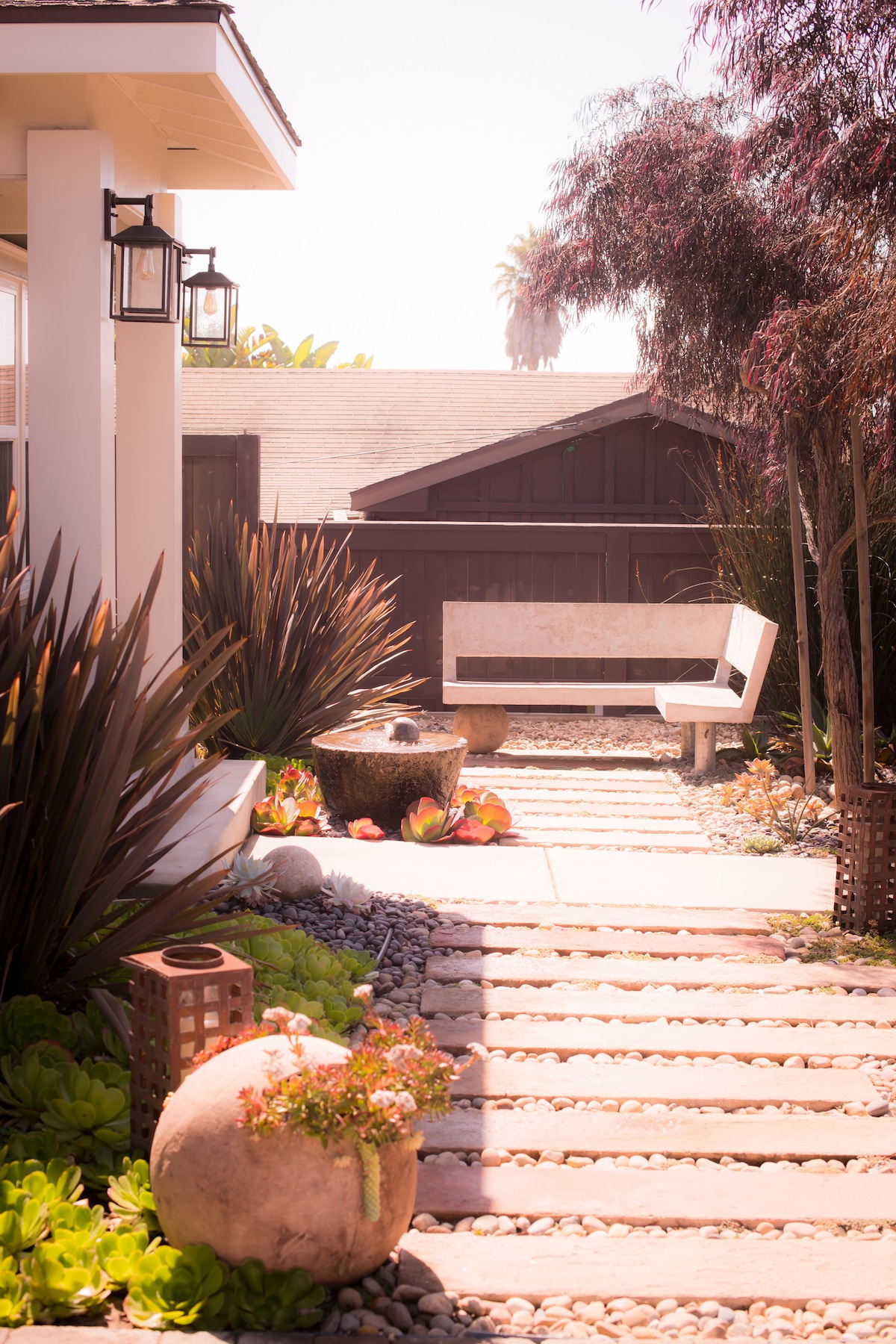 A well-maintained pathway leads through landscaped gardens featuring a variety of succulents and decorative rocks. An elegant bench sits alongside the path, with stylish lanterns hanging above. The surrounding area is framed by lush foliage, creating a serene outdoor ambiance.