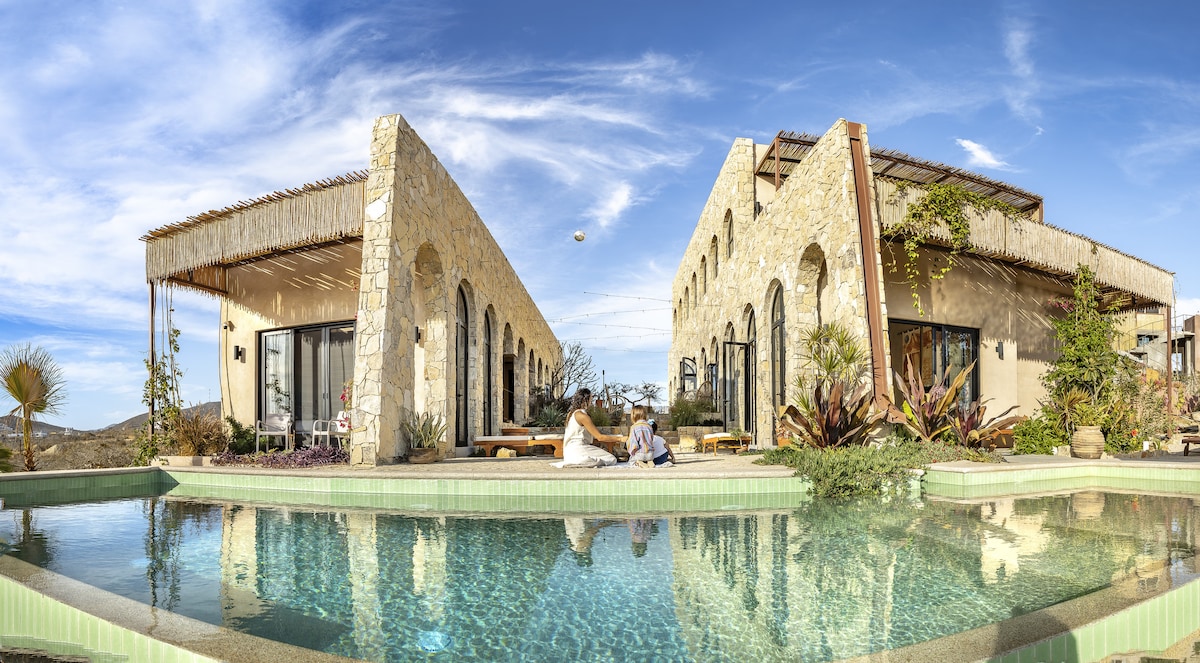 A panoramic view of a luxury villa is showcased, highlighting two elegantly designed structures flanking a serene oasis pool. Lush landscaping surrounds the pool, with clear skies and distant hills contributing to the tranquil atmosphere. Open spaces invite natural light into the cheerful settings.
