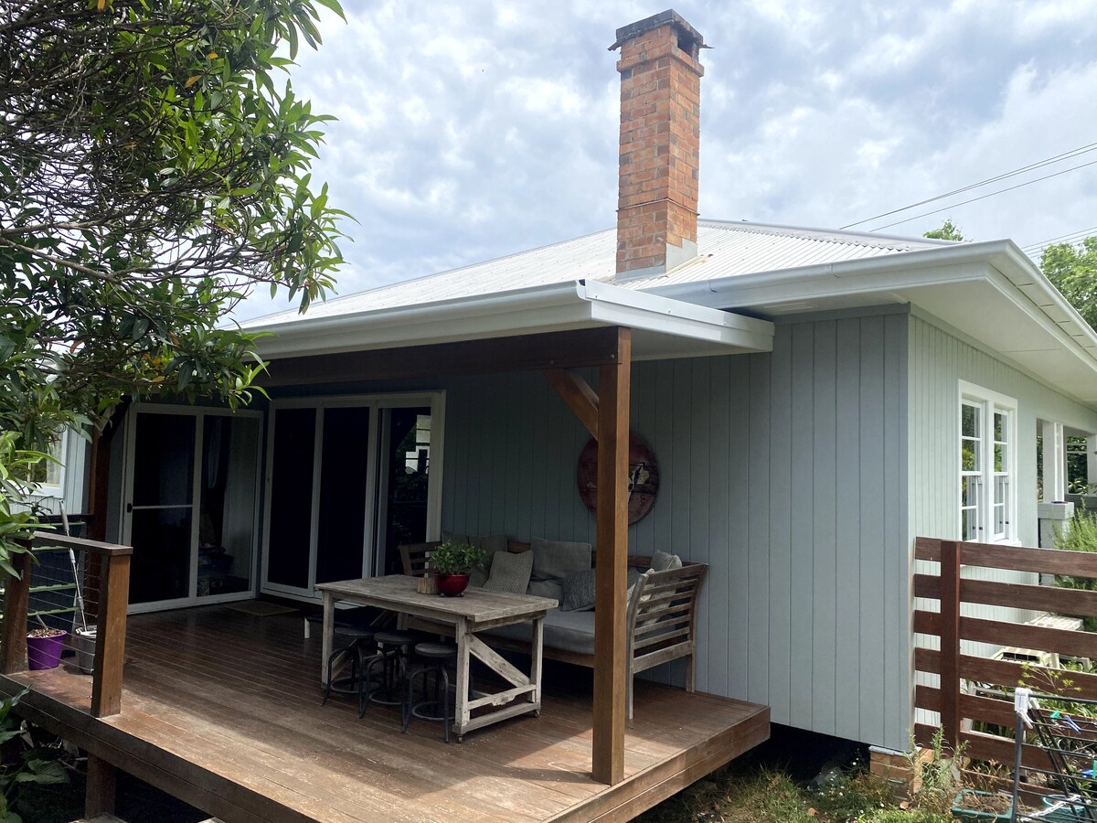 A covered deck area features a wooden table and chairs, providing a comfortable outdoor space. Potted plants add touches of greenery, while the exterior of the light-colored cottage showcases a traditional chimney and large windows that allow natural light inside.