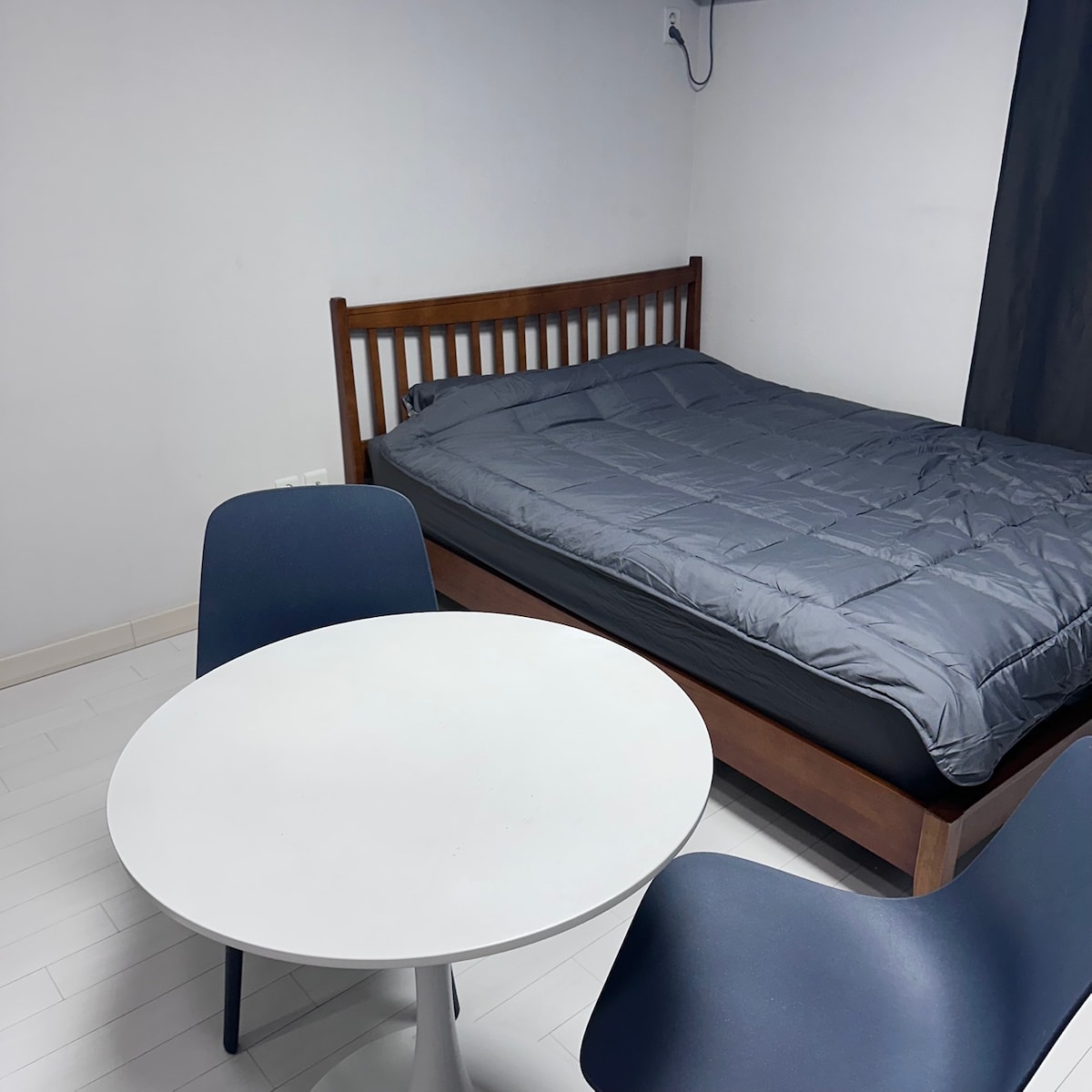 A simple room features a wooden bed with a dark comforter. A round white table occupies the center, accompanied by two blue chairs. Light walls and a dark curtain contribute to the room's understated aesthetic.