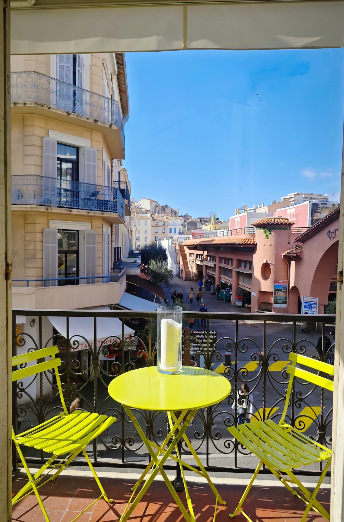 A sunny terrace is depicted, featuring a round yellow table accompanied by two matching chairs. The view showcases a quiet pedestrian street, with buildings lining the sides and clear blue skies above, creating an inviting outdoor space.