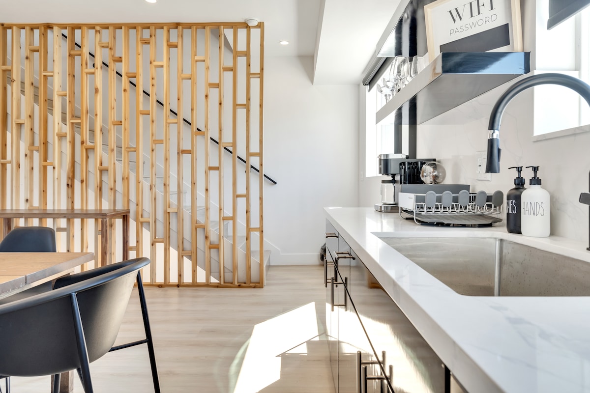A modern kitchen space features a sleek countertop and a neatly organized arrangement of kitchenware. Natural light enters through large windows, accentuating the light wood finishes and the decorative wooden partition in the background, which complements the minimalist interior design.