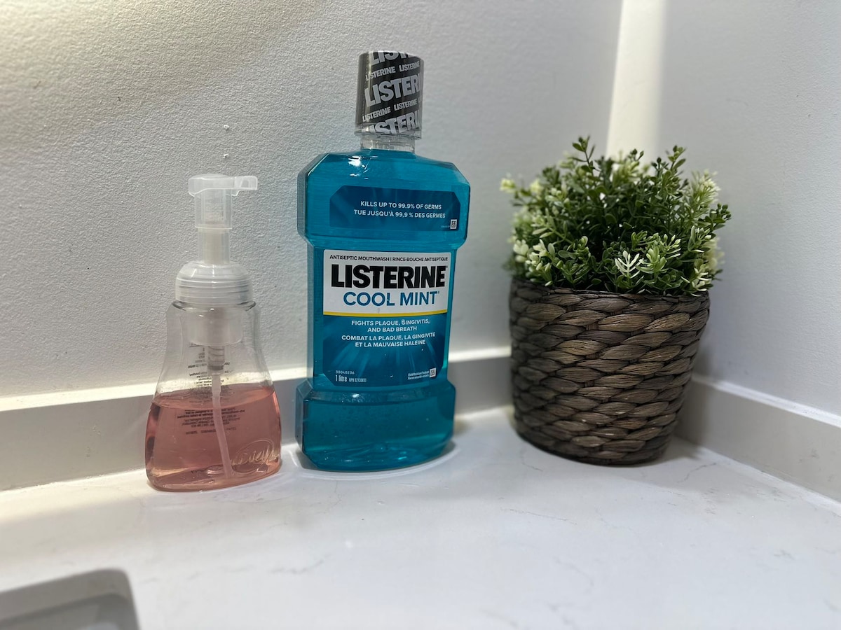 A bathroom counter displays a bottle of blue mouthwash beside a pink liquid soap dispenser. A small, woven planter containing greenery sits nearby, contributing to the neat and organized appearance of the space.