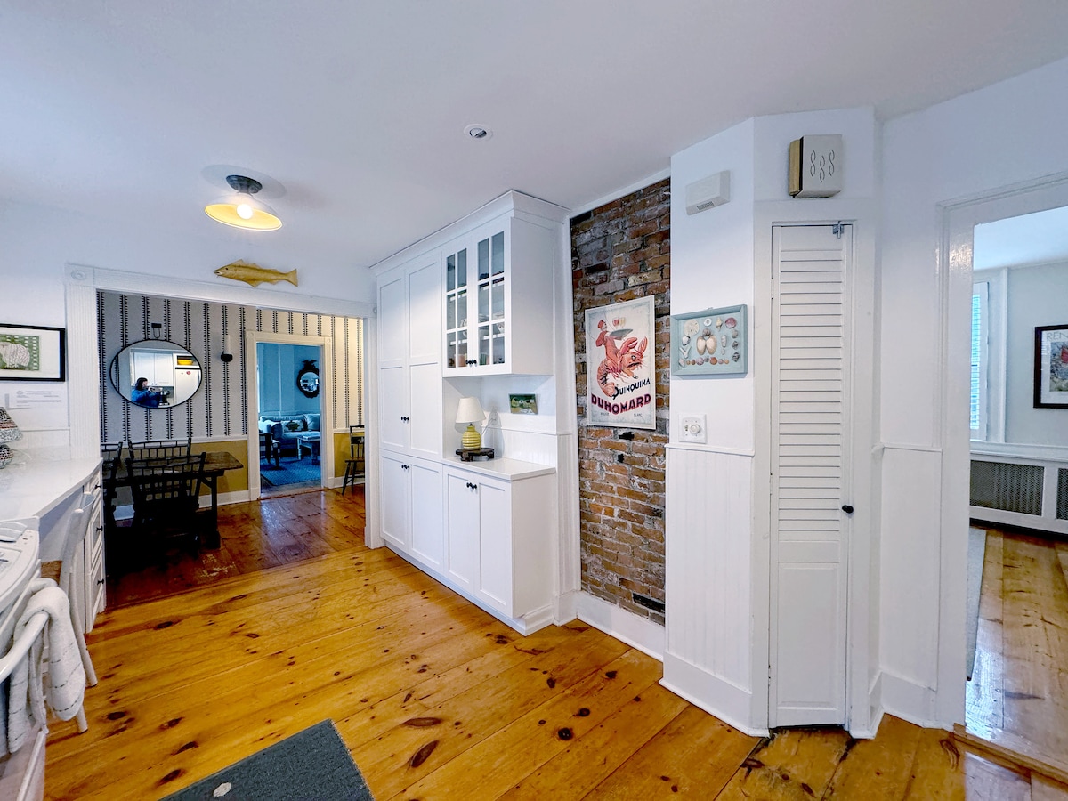 The kitchen area features white cabinetry and a decorative brick wall, creating a charming contrast. A small round dining table is visible in the background, and light wood flooring adds warmth to the space. A wall with framed art enhances the inviting atmosphere.