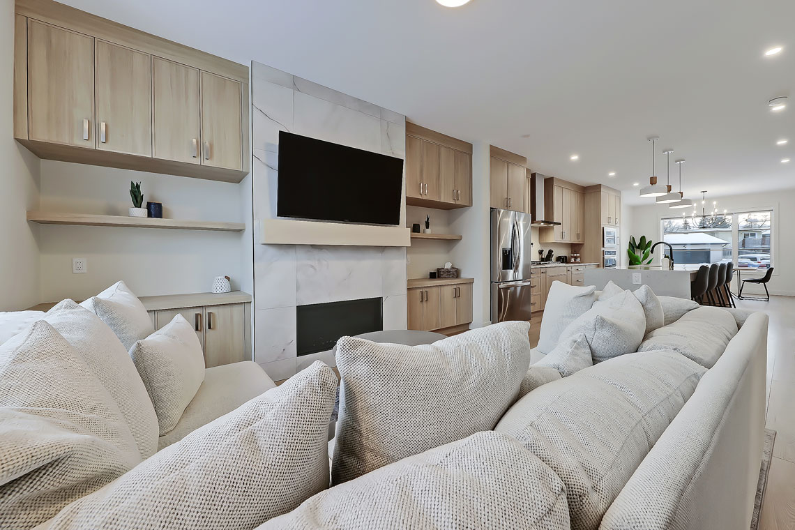 The open-concept main living area features a plush sectional sofa dressed with soft, textured pillows. A modern electric fireplace adds a subtle warmth, while a large flat-screen TV is mounted above a sleek stone accent wall. Natural light streams in from the adjacent kitchen.
