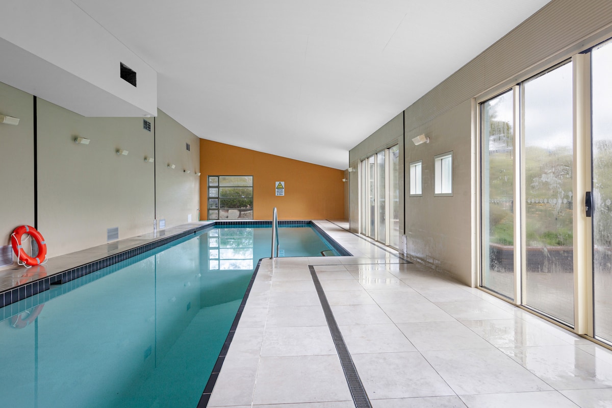 An indoor heated pool area is showcased with large windows allowing natural light to fill the space. The pool features a smooth tiled edge and safety equipment is visible, providing a clean and inviting environment for swimming and relaxation.