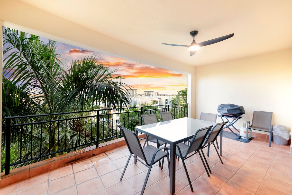 An outdoor balcony features a modern dining table with six chairs, surrounded by greenery. A BBQ grill is positioned nearby, and a ceiling fan provides airflow. The vibrant sunset colors reflect off the horizon, enhancing the serene atmosphere of this outdoor space.