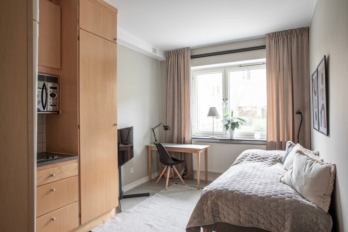 A studio layout is shown, featuring a well-equipped kitchen area with wooden cabinetry. A comfortable sleeping area includes a spacious bed and a work desk positioned by large windows that allow natural light. Soft curtains complement the neutral color scheme.