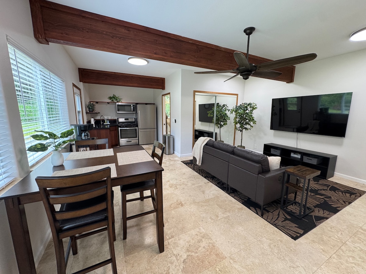 An open living space features a dark sofa facing a mounted television against a light wall. A dining table for four is situated near large windows providing natural light. Kitchen appliances are visible in the background, complemented by natural wood beams overhead.