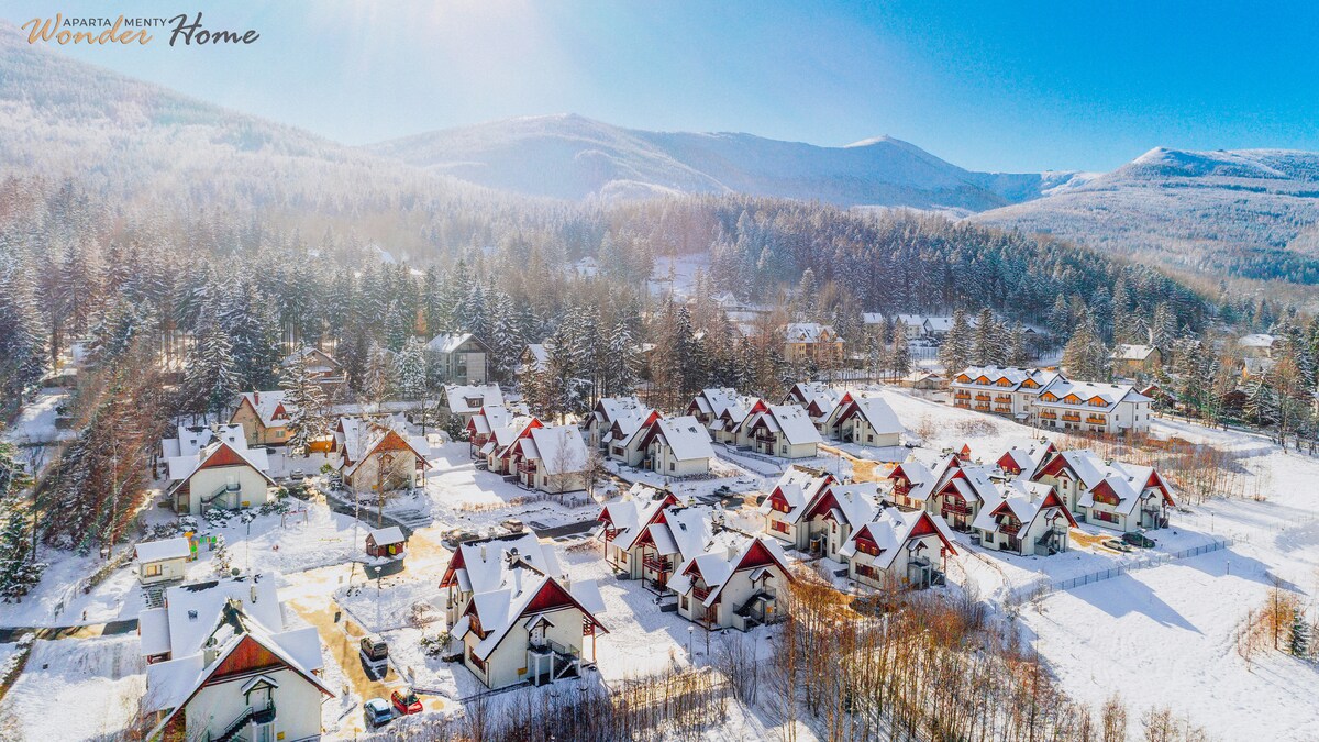 A scenic aerial view captures a winter landscape blanketed in snow, showcasing a cluster of chalets with red roofs. Surrounding mountains create a serene backdrop, while the area is marked by trees dusted with snow, conveying a tranquil atmosphere.