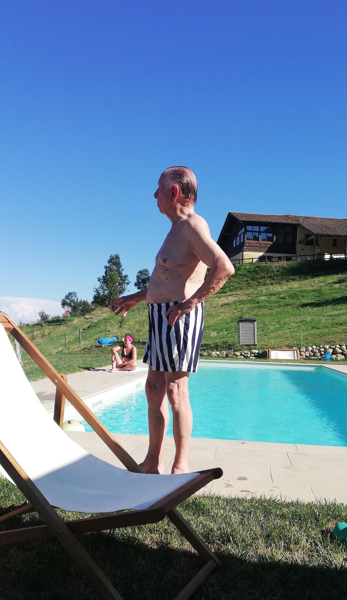 A serene poolside scene presents a swimming pool surrounded by well-maintained grass. A sun lounger is positioned nearby, and a two-story building is visible in the background, set against a clear blue sky. The setting offers a peaceful view of the adjoining hills.