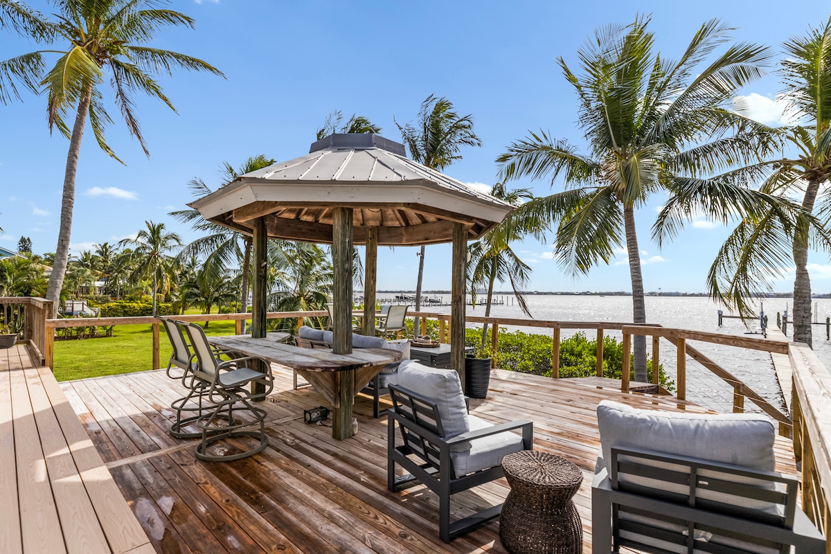 A spacious outdoor deck features a gazebo, surrounded by palm trees and offering views of the water. Comfortable seating arrangements include lounge chairs and a dining table, creating an inviting space for relaxation and enjoyment of the serene surroundings.