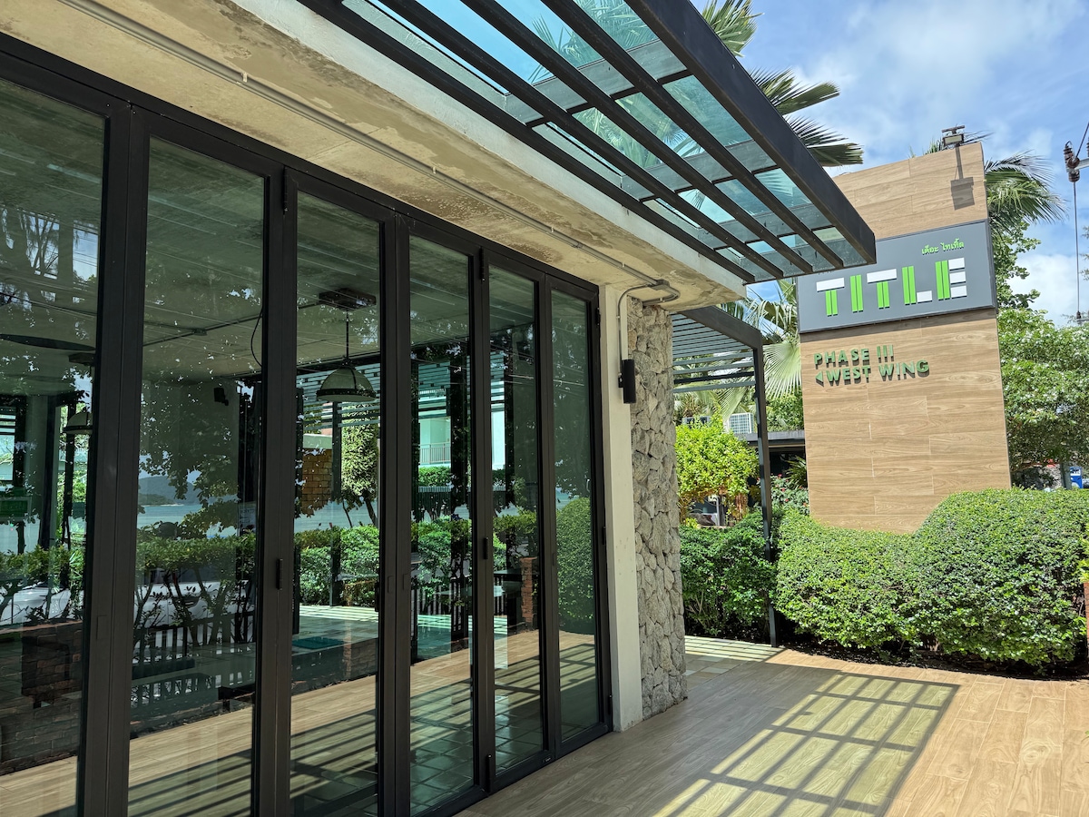 The image shows a modern building with large glass doors and a covered entrance. Surrounding greenery adds a lush ambiance, while a sign with the word 'TITLE' is visible on the adjacent structure. A wooden deck provides an inviting access point.