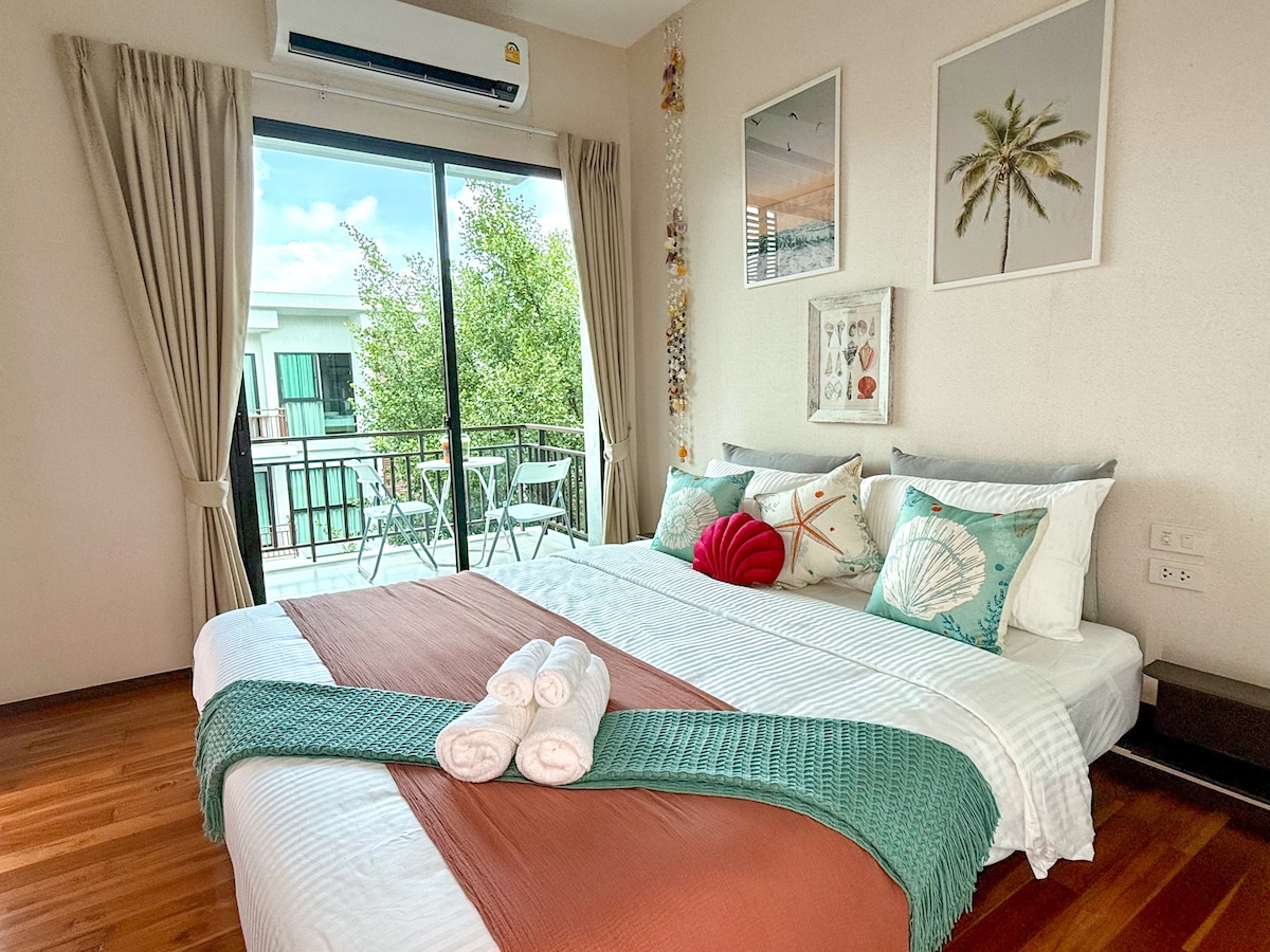 A bright sleeping area features a spacious bed adorned with decorative pillows and neatly rolled towels. A large window opens to a balcony, allowing natural light to fill the room. Stylish wall art and warm wood flooring enhance the tranquil setting.