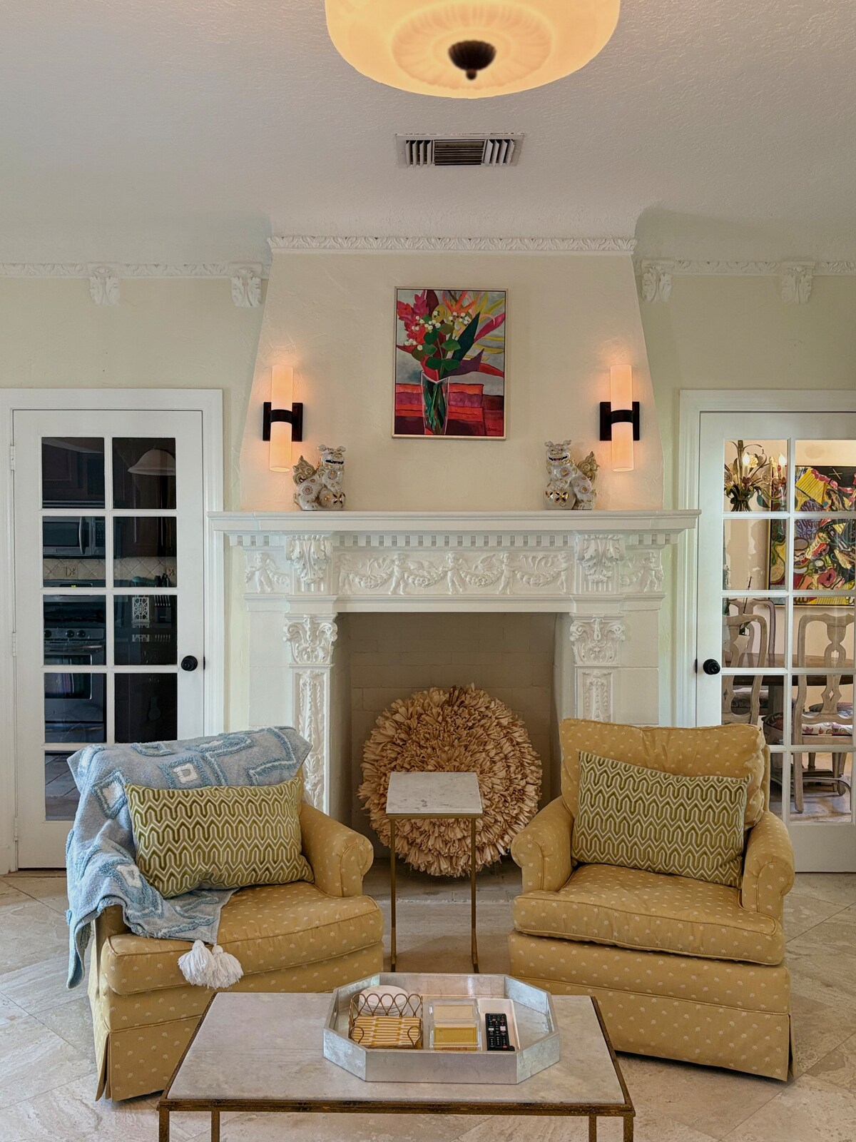 This inviting living room features a detailed fireplace, soft lighting, and colorful artwork. French doors and classic trim add charm, while thoughtful decor and cozy touches create a warm, relaxing space filled with natural light.