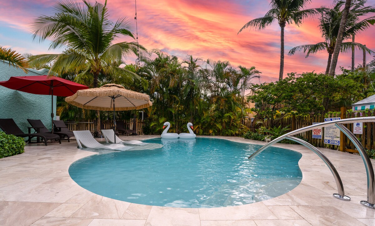 A serene heated saline pool is surrounded by lush greenery and palm trees, with a beige stone deck offering lounge chairs and umbrellas for shade. Two white swan-shaped floats rest gently in the water, enhancing the tranquil ambiance during sunset.