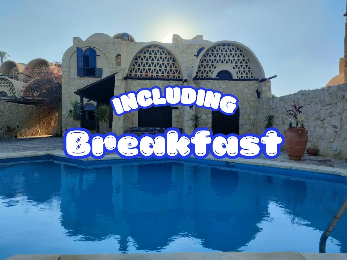 A serene swimming pool is showcased, reflecting the clear blue sky above. The surrounding stone wall features unique arch designs, adding character to the outdoor space. An inviting area for relaxation is created with ample sunlight filtering down, enhancing the tranquil atmosphere.