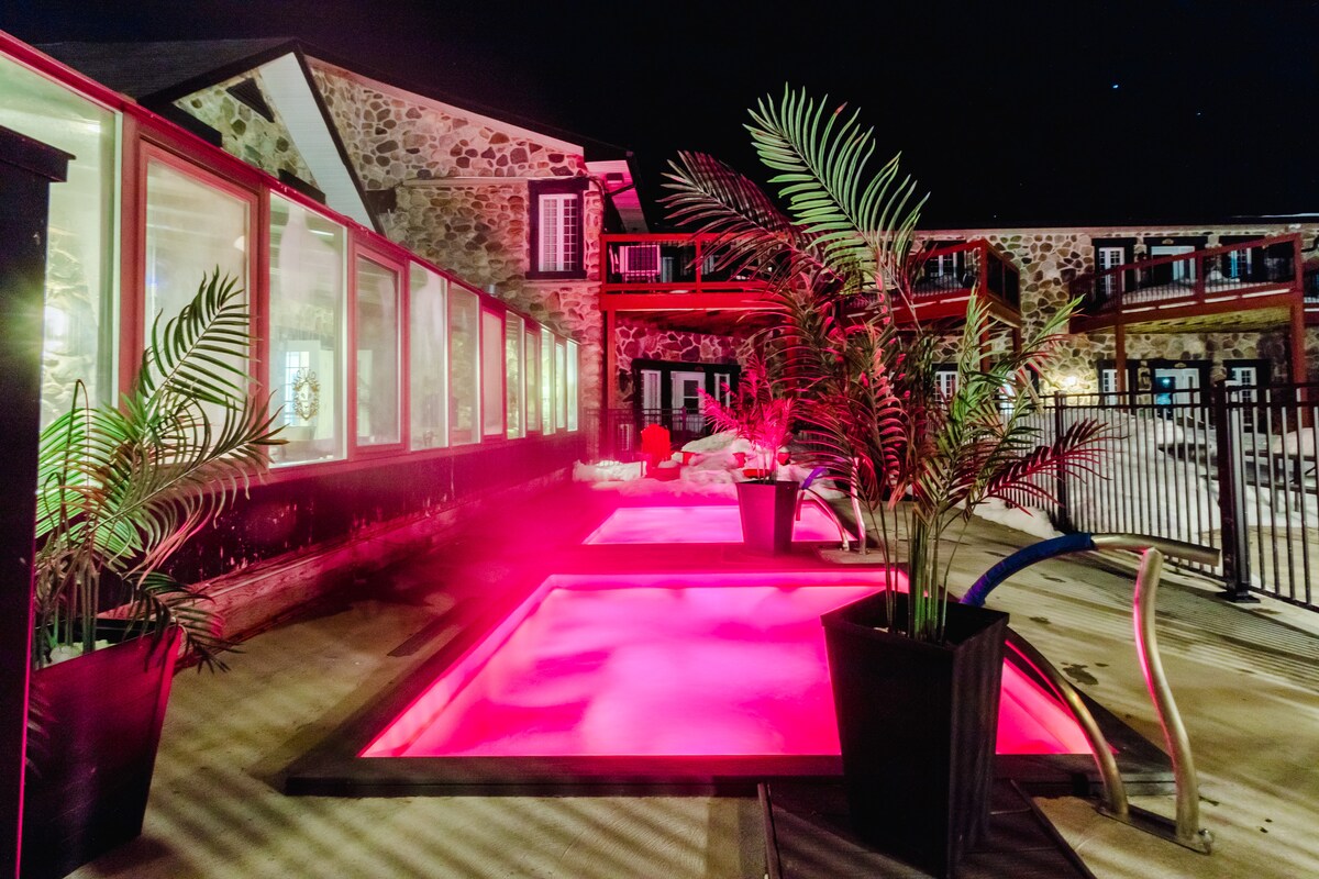 The outdoor area features illuminated spas framed by tropical plants in large pots. The soft glow of pink lighting highlights the textured stone wall of the historic building nearby, while the surrounding patio provides ample space for relaxation.