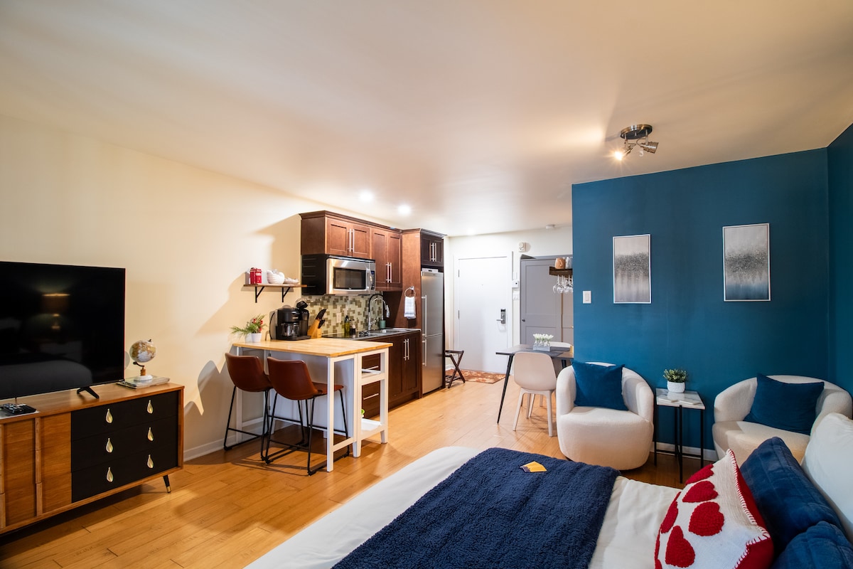 An open living space features a cozy sleeping area with a spacious bed dressed in navy bedding. A dining table with two chairs is positioned beside a kitchenette equipped with modern appliances. Light wood flooring complements the dark cabinetry, and a television is placed on an accent console.