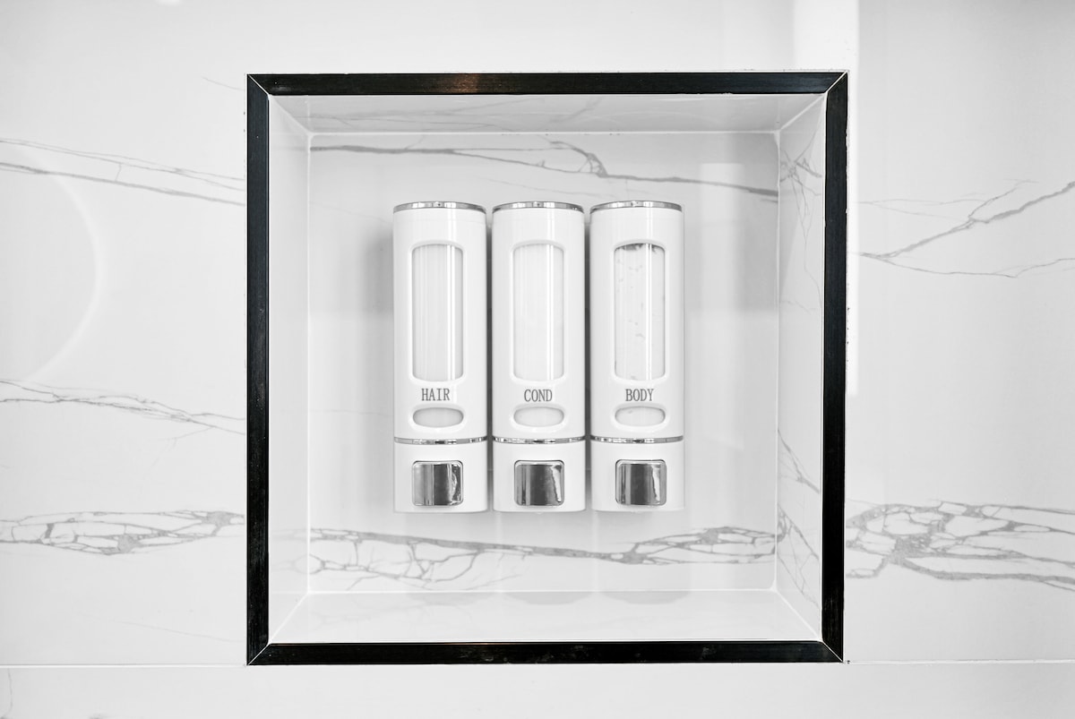 Three sleek dispensers are mounted on a modern, marble-patterned wall. Each container is labeled for hair, conditioner, and body wash, providing convenience in the bathroom. The minimalist design enhances the space's contemporary aesthetic.