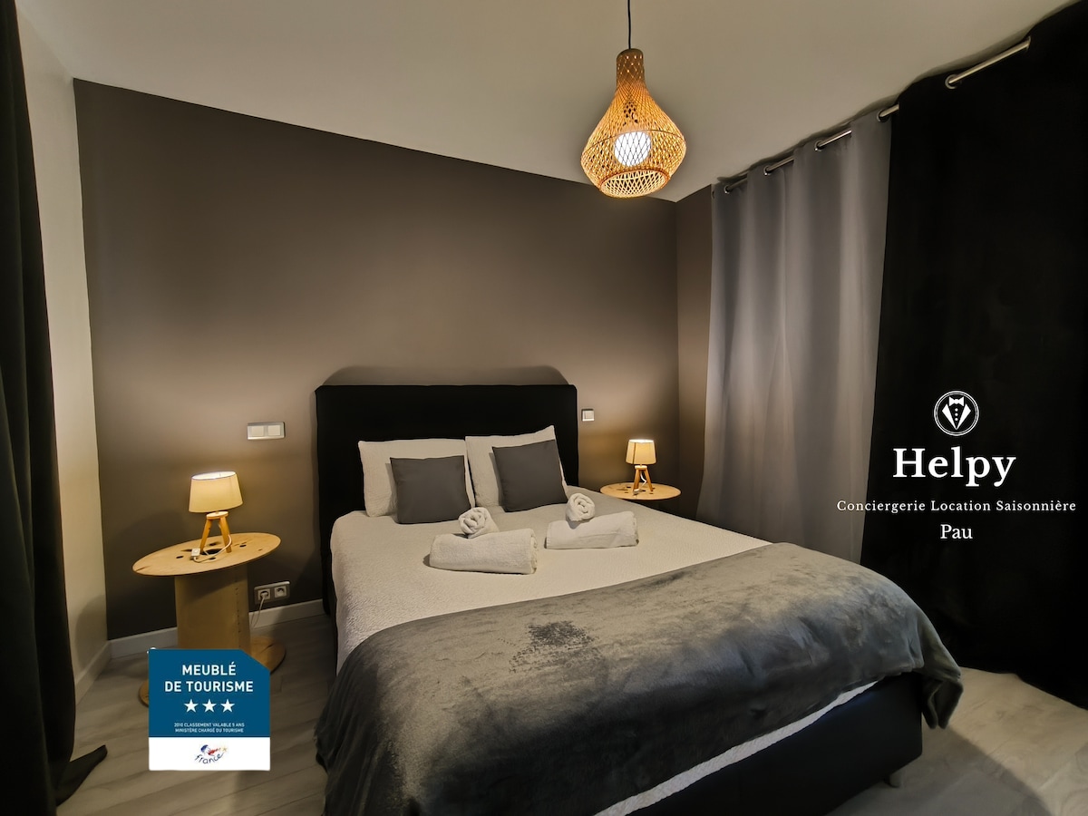 A serene bedroom features a spacious bed with neatly arranged linens and towels. The room is complemented by two bedside tables with soft-lit lamps. Dark curtains provide privacy, while a decorative woven light fixture adds a warm touch to the ambiance.