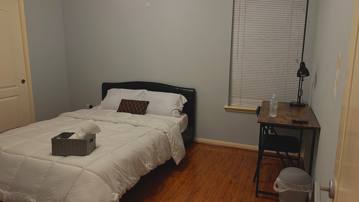A spacious bedroom features a comfortable king-size bed with a light-colored bedding set. A small desk with a chair is positioned nearby, accompanied by a lamp. Natural light filters in through a window with blinds, and the warm wooden floor adds a cozy feel to the room.