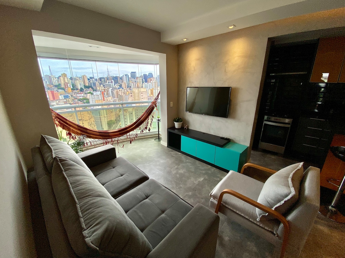 A modern living area is viewed from an angle, featuring a spacious gray sofa and a light-colored lounge chair. A mounted flat-screen TV is positioned above a teal media console. The floor-to-ceiling windows provide a sweeping view of the São Paulo skyline.