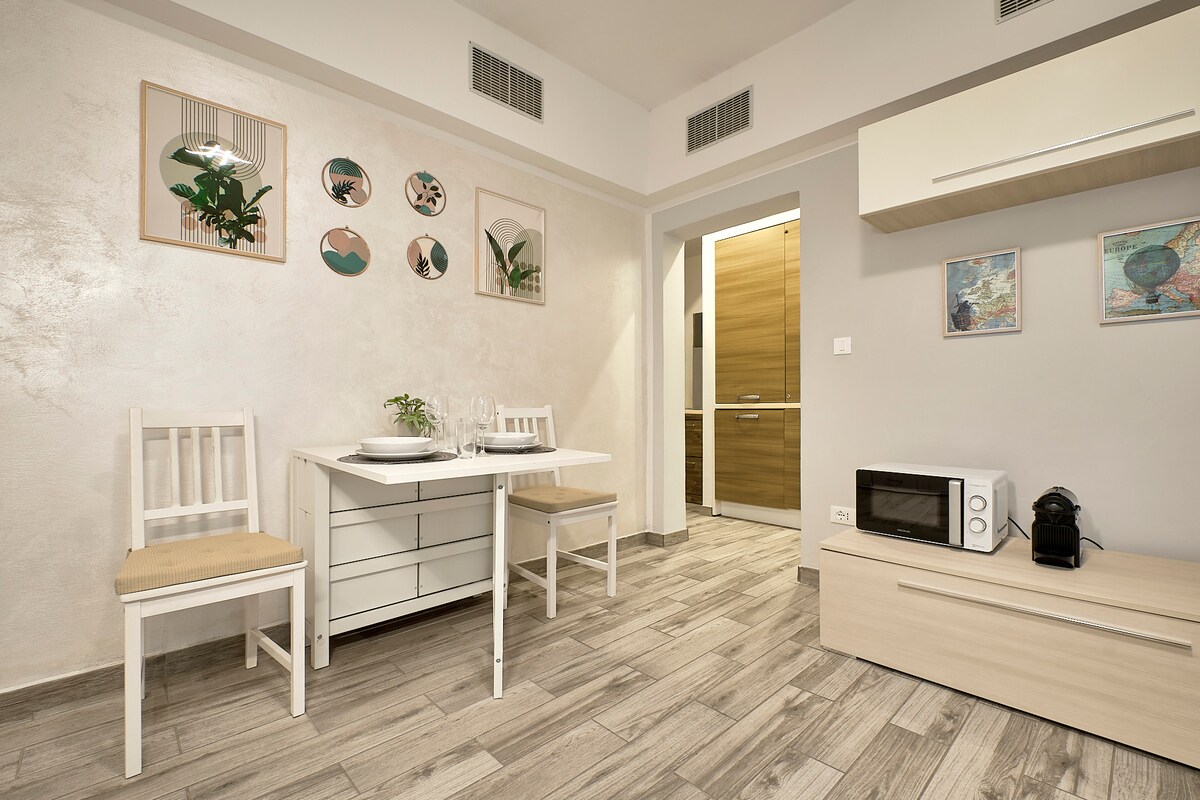 A bright dining area features a simple white table set for two, accompanied by matching chairs. On the wall, artistic prints adorn the space, while a compact kitchen area is visible with a microwave and a coffee machine nearby. The floor is tiled with light wood patterns.