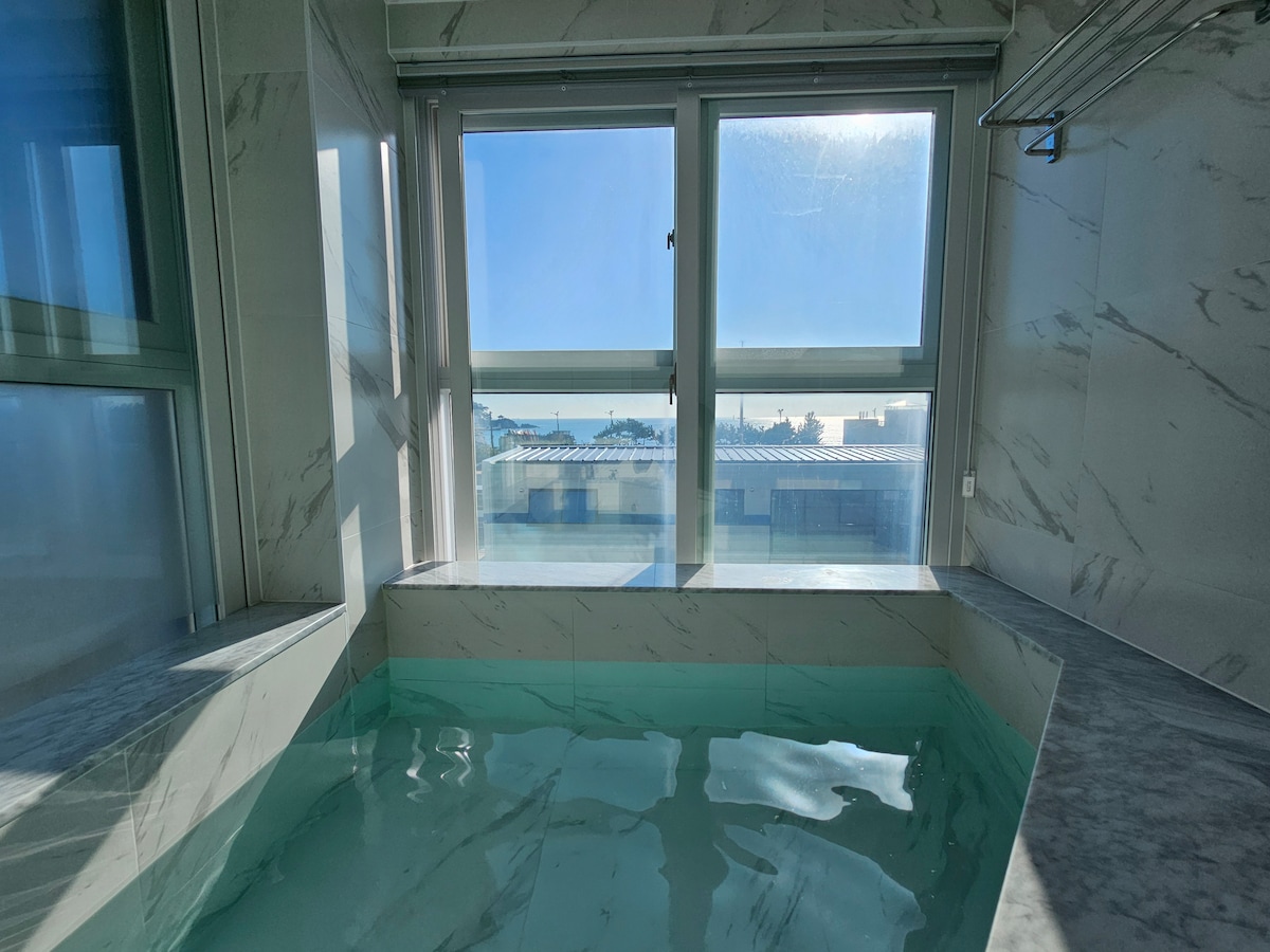 The image displays a jacuzzi set within a well-lit room. Large windows allow bright natural light to illuminate the space, showcasing the relaxing water surface. The marble design of the surrounding walls enhances the contemporary feel of the room.