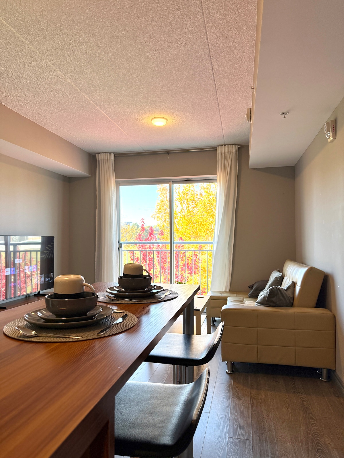 A dining area features a polished wooden table set with plates and candles. A comfortable leather sofa and a small cushion are arranged next to a large window, showcasing vibrant fall foliage outside. Natural light filters through soft curtains.
