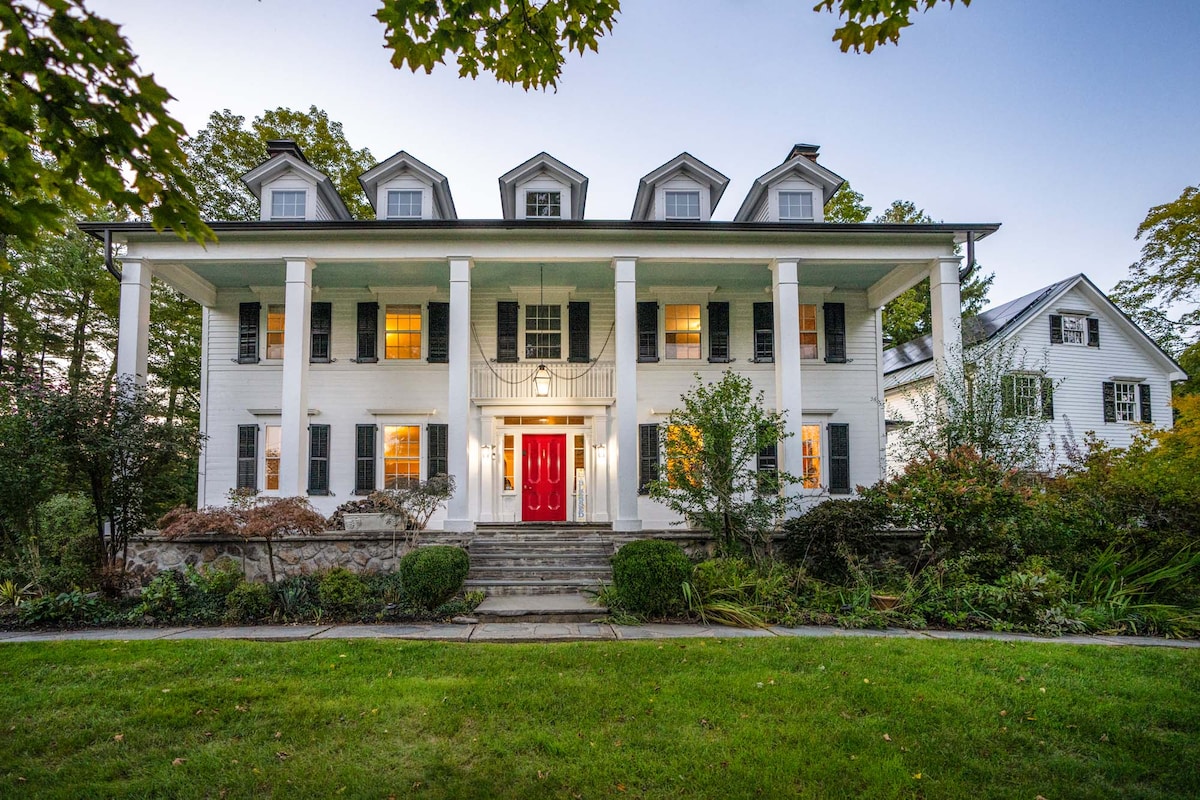 The historic estate features a grand facade highlighted by multiple gabled roofs and a striking red front door. Soft lighting glows from the windows, and lush greenery surrounds the stone steps leading to the entrance, emphasizing the peaceful atmosphere of the property.