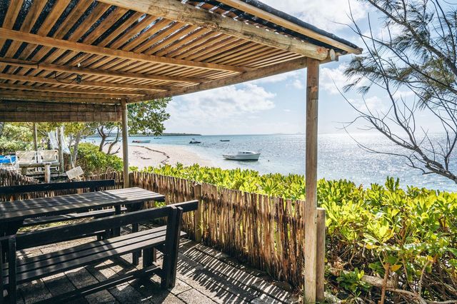 A shaded outdoor area features dark wooden tables and benches set against a natural backdrop. The sandy beach extends into the distance, met by tranquil waters reflecting the sky. Lush greenery frames the scene, offering a serene and inviting atmosphere for relaxation.